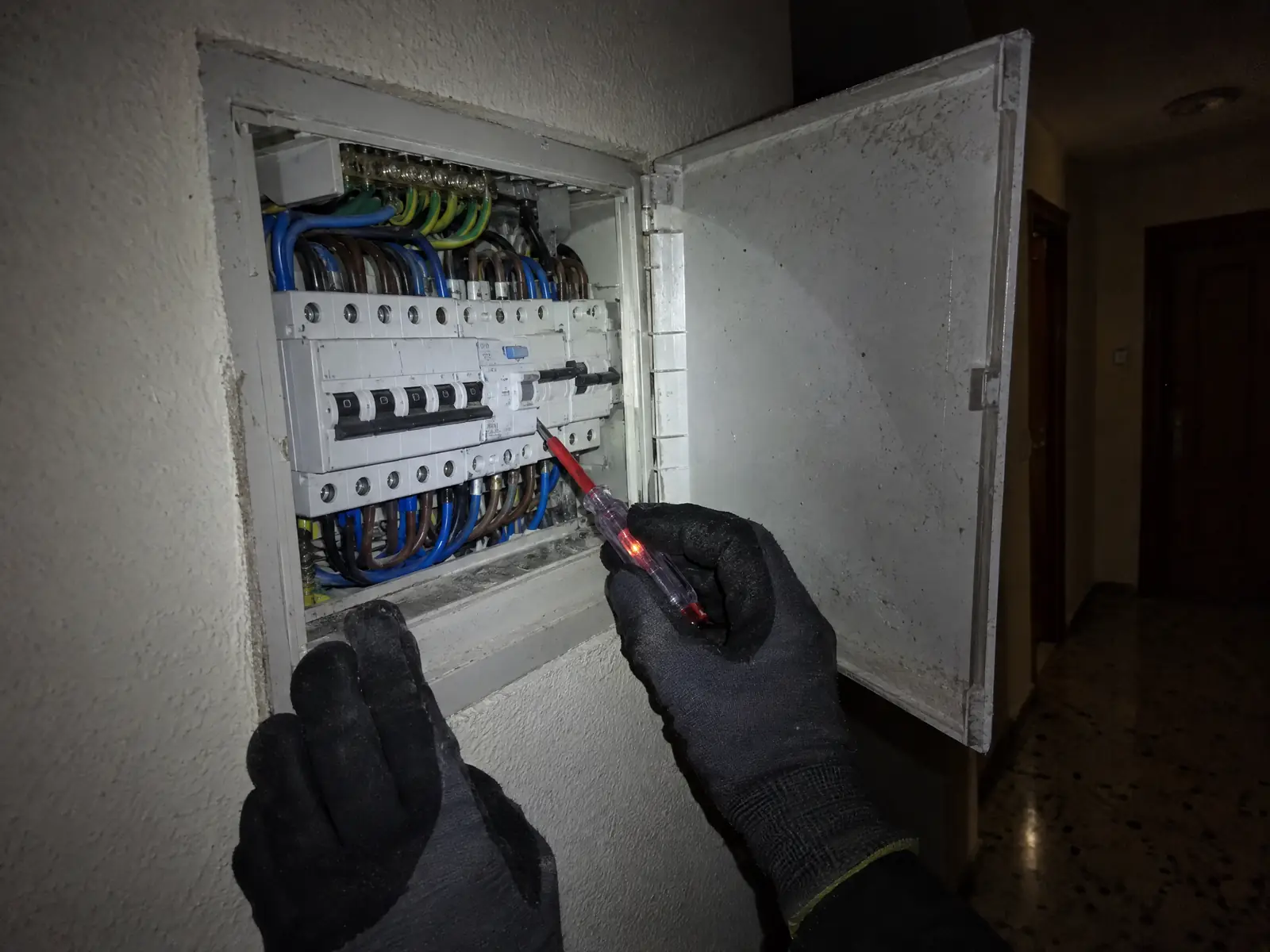 Manos con guantes comprueban con un buscapolos un cuadro eléctrico abierto en el pasillo de un piso, a oscuras y con luz de linterna