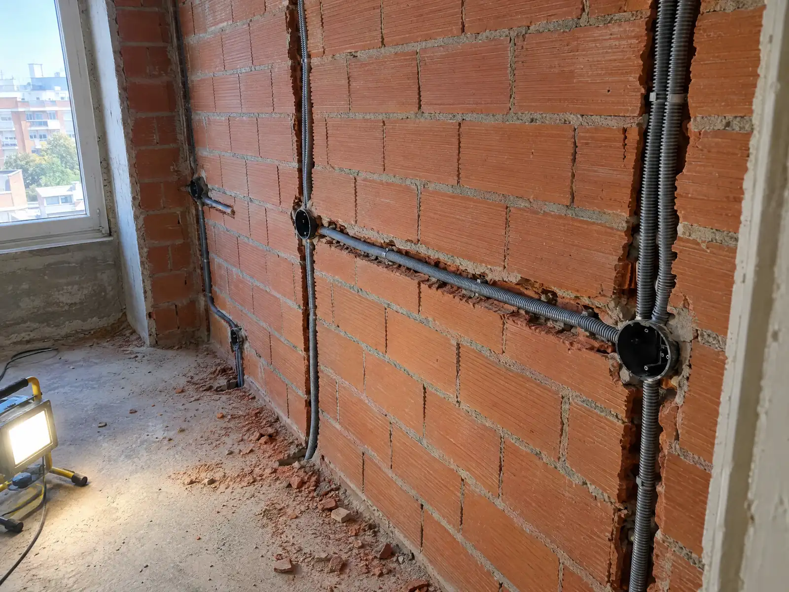 Pared de ladrillo con rozas recién abiertas, tubo corrugado gris tendido hasta las cajas y escombro en el suelo